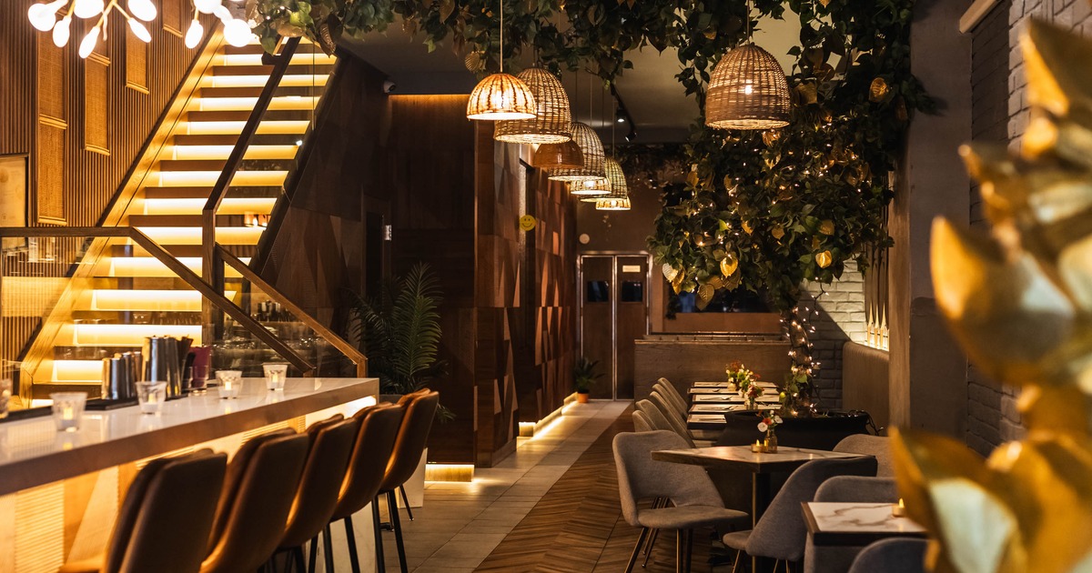 Cozy restaurant with warm lighting, modern bar with high stools, lush hanging plants, wooden decor