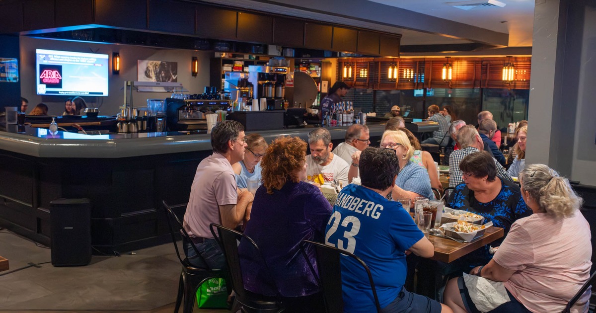 A group of people dine and socialize at tables in the modern restaurant with a prominent bar
