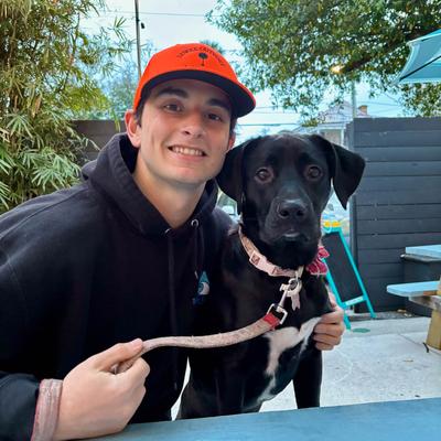 Smiling person in an orange cap and black hoodie sits next to a calm black dog outside.