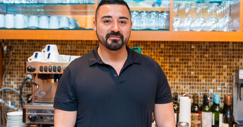A headshot of an employee,  with cups, glasses, and a coffee machine in the background