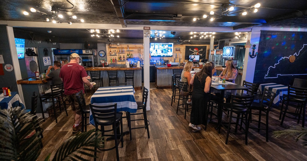 A cozy bar with wooden floors and string lights, featuring a central bar counter and patrons seated at tables