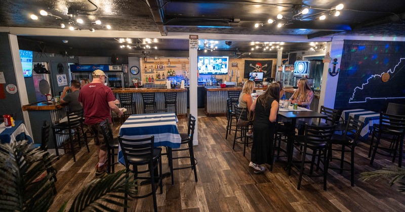 A cozy bar with wooden floors and string lights, featuring a central bar counter and patrons seated at tables
