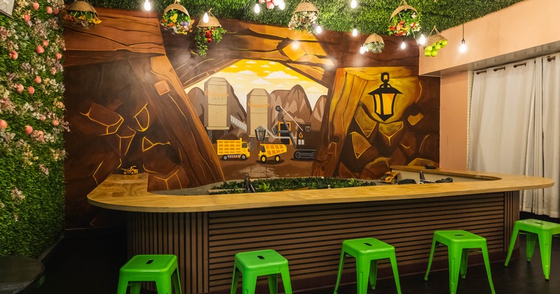 Indoor space with green foliage on ceiling, mural on wall, wooden counter, and green stools