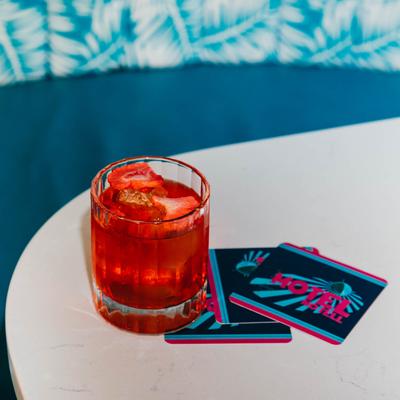 A vibrant red cocktail garnished with fruit, sits on a table, blue seating is in the background.