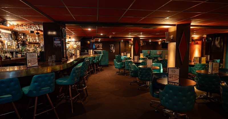 Interior of a stylish bar featuring a long curved bar counter, turquoise seating, and dim lighting