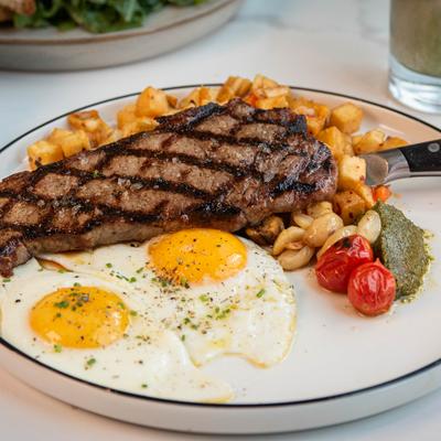 Grilled steak, sunny-side-up eggs, breakfast potatoes, and tomatoes on a white plate.