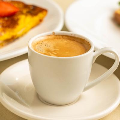 A small white cup and saucer containing a shot of espresso, food plates in the background.