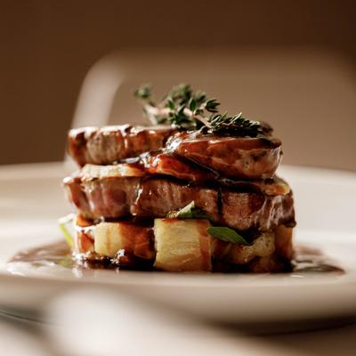 A plate of three stacked slices of meat with sauce, roasted vegetable cubes and herbs on top.
