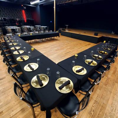 Dining set up with a U-shaped table arrangement featuring black tablecloths and gold plates.