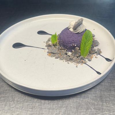 A round purple dessert sits on a white plate, garnished with mint leaves and whipped cream.