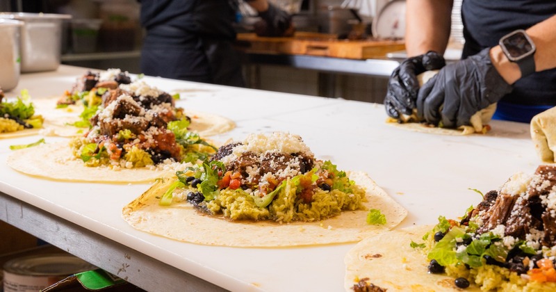 Chefs in a kitchen preparing burritos, filling tortillas with rice, guacamole, beans, and cheese