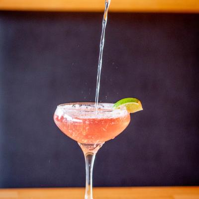 Cosmopolitan cocktail being strained into a coupe glass with a lime wedge garnish.