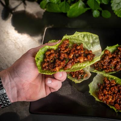 A person holds a lettuce wrap with minced meat filling above the plate with more wraps.