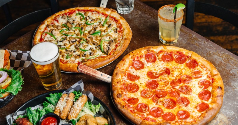Two pizzas on a table with a platter of fried foods, beer and cocktails