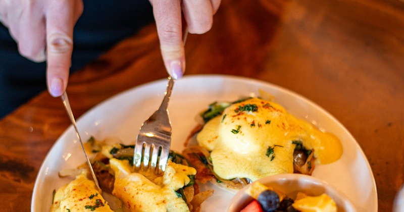 Person cutting into a Eggs Benedict served with a bowl of fruits