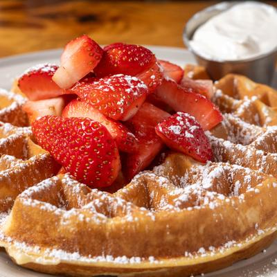 Waffle topped with powdered sugar and fresh strawberries, a side of whipped cream.