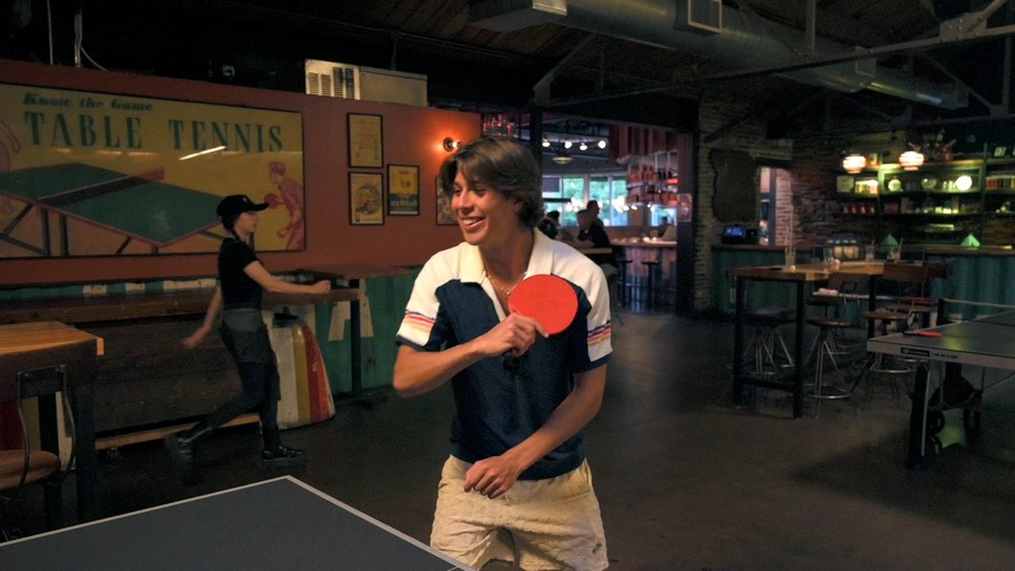 Interior, a customer holds a ping pong paddle by the table during a game.