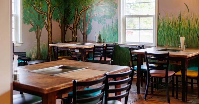 Restaurant interior with wooden tables, inset grills, and a forest-themed mural