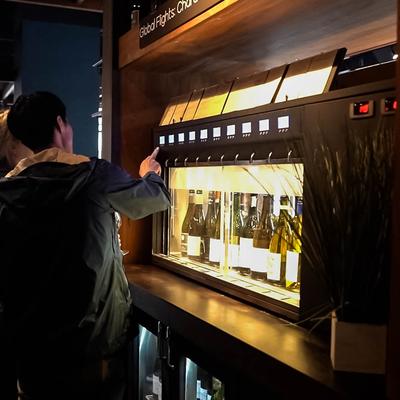 A person interacts with a lit wine dispenser displaying several bottles.
