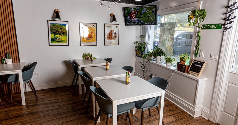 A modern cafe corner with white tables, gray chairs, wall art, ceiling lights, and TV