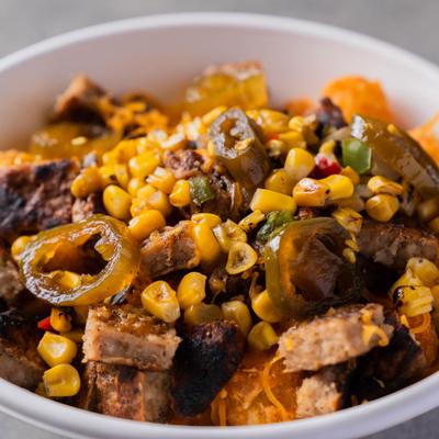 Close-up of a bowl filled with grilled meat, corn, sliced peppers & roasted vegetables.