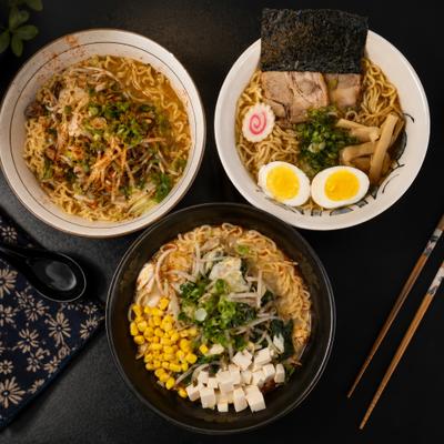Three bowls of diverse ramen sit on a dark table with chopsticks.