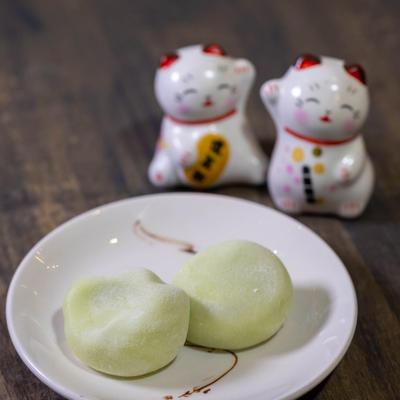Two green mochi pieces on a plate with lucky cat figurines in the background.