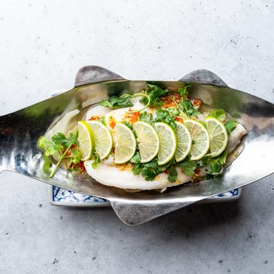 Steamed fish topped with lime slices, cilantro, and chili in a metal dish.