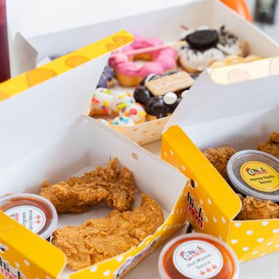 Fried chicken tenders in yellow boxes with dipping sauces and background donuts.