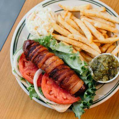 A bacon-wrapped hot dog with lettuce, tomato, and onion, served with fries and coleslaw