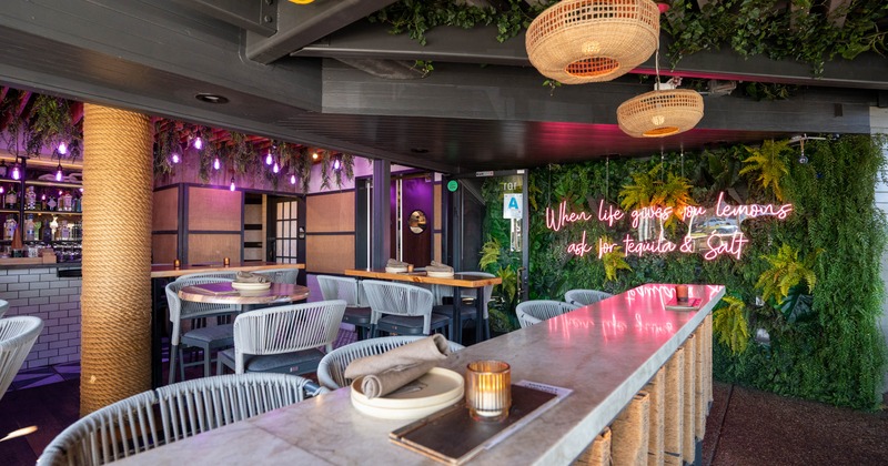Colorful interior, marble bar, woven seating, hanging lights, leafy wall displaying a pink neon sign