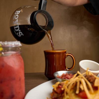 A person pouring coffee into a mug on a table with served tacos and Bloody Mary.