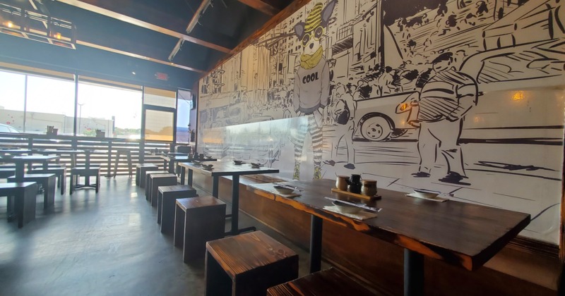 Spacious restaurant with wooden tables and benches, featuring a large, playful comic-style wall mura