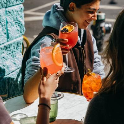 Three people at an outdoor table raise orange cocktails in a toast.