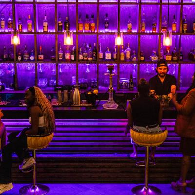 Bar area featuring a back bar illuminated by purple lighting and shelves stocked with various bottles.