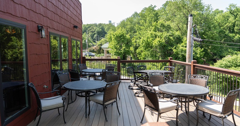 Wooden deck - metal tables and chairs, next to a red building, overlooking a green wooded area