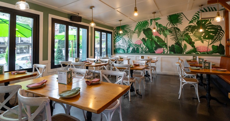 A restaurant interior with tropical mural, wooden tables, and white chairs
