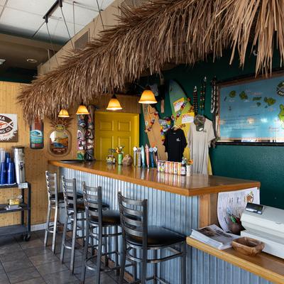 Bar interior with a thatched roof, wooden counter, bar stools, surfboards and Hawaiian decor.