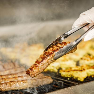 Grilled kebab being flipped with kitchen tongs, smoke rising from the barbecue grill.