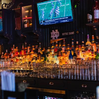 Bar area featuring liquor bottles on backlit tiered shelves and an overhead TV showing sports.
