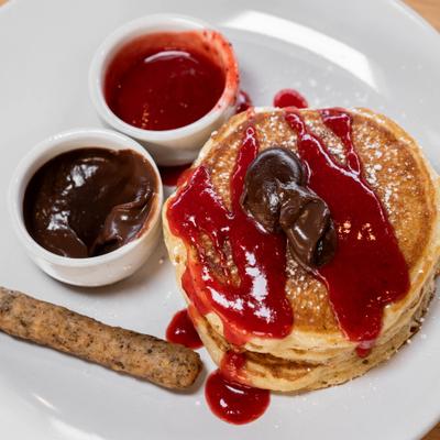 A stack of pancakes topped with a berry compote, chocolate ganache, and a sausage link.
