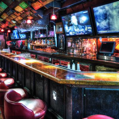 A wooden bar counter with red stools, overhead glasses, liquor bottles, and glowing TV screens