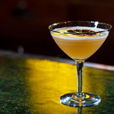 Golden cocktail in coupe glass topped with dried citrus slice on bar.