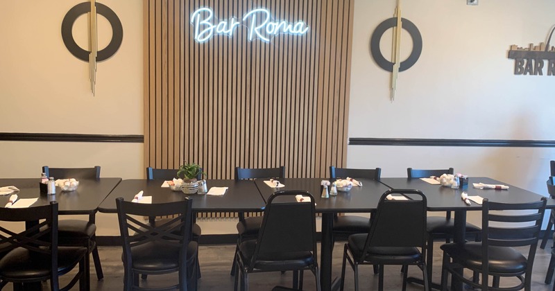 Restaurant interior with black tables, chairs, a neon "Bar Roma" sign and circular wall art.