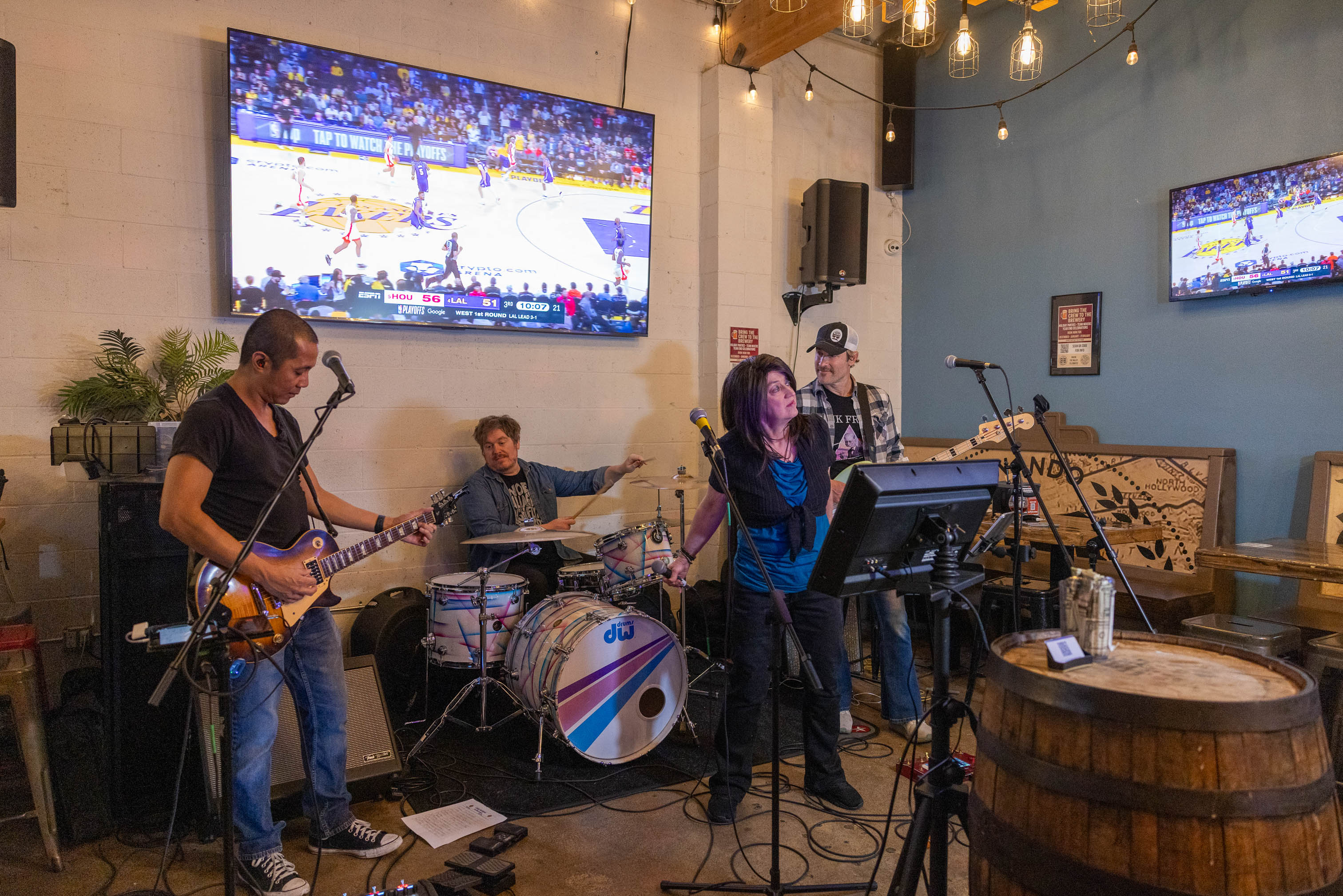 Live band performing on a small stage with an overhead TV showing a basketball game