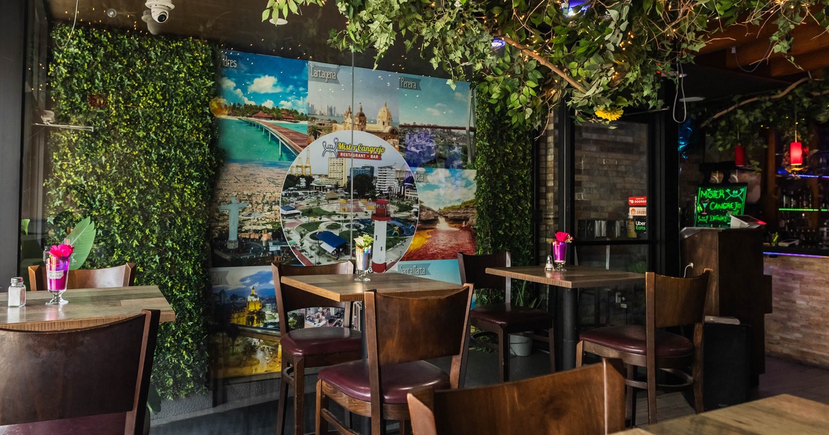 Interior of a restaurant with wooden tables and chairs, green foliage, a mural on the wall