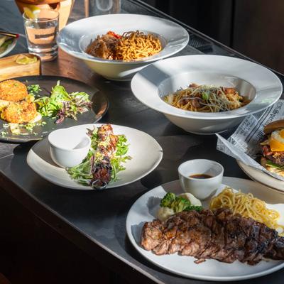 A variety of dishes on a metal table, including pasta, a burger with fries, skewers, and crabcakes.