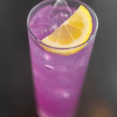 Purple Lemonade served in a highball over ice, garnished with a lemon slice.