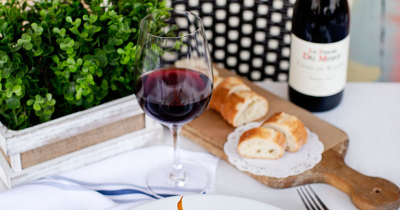A glass of wine on a table with greenery, bread and a bottle of red wine