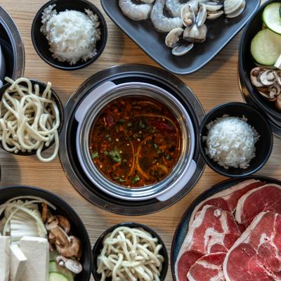 Broth in a pot alongside cups of white rice and noodles, meat and shrimp plates.